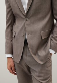 Abito marrone chiaro strutturato con chiusura a due bottoni, rever a intaglio e tasche laterali, abbinato a una camicia bianca e una cintura nera.
