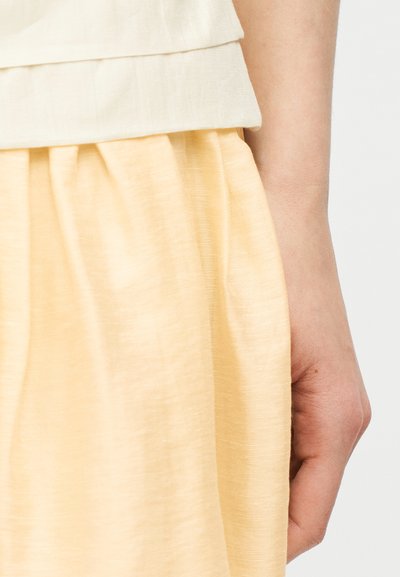 Δεξί χέρι ακουμπισμένο στο πλάι, φοράει φούστα σε ανοιχτό κίτρινο χρώμα και τοπ με στρώσεις σε εκρού, πάνω σε απλή ανοιχτόχρωμη βάση.