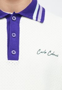 Biała koszulka polo z krótkim rękawem wykonana z teksturowanej tkaniny, z fioletowym paskowanym kołnierzykiem i dwoma fioletowymi guzikami. Na niej wyhaftowane na zielono "Carlo Colombo".