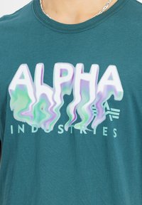 Tyrkysové tričko s krátkým rukávem, které má velký, deformovaný grafický nápis "ALPHA" ve odstínech bílé, zelené a fialové, s "INDUSTRIES" pod ním.