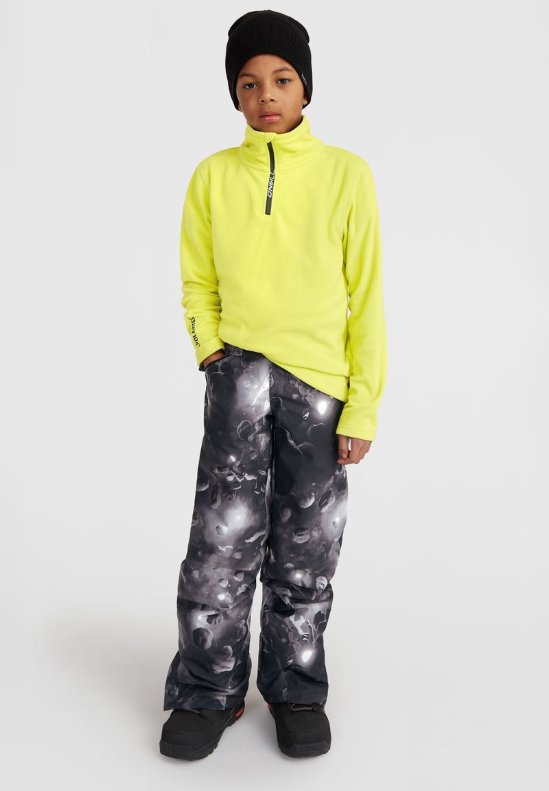 Pull en polaire jaune vif avec zip quart, associé à un pantalon de ski noir et gris imprimé avec un motif texturé. Bonnet noir et bottes.