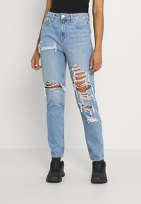 Jeans en denim bleu clair avec des détails déchirés et des bords effilochés. Caractérisés par une coupe décontractée et une poche zippée sur le côté. Portés avec des baskets noires.