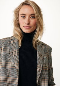 Blazer con motivo a scacchi in marrone, nero e bianco, indossato su un dolcevita a coste nero. Texture morbida, vestibilità su misura.