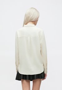 Camisa de manga longa em creme claro, com colarinho, tecido texturizado, pala nas costas e punhos abotoados, combinada com uma saia preta evasê.