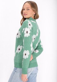 Grüner Strickpullover mit weißen Blumenmustern und schwarzen Zentren. Gerippte Bündchen und Saum. Weiche Textur mit lockerer Passform.
