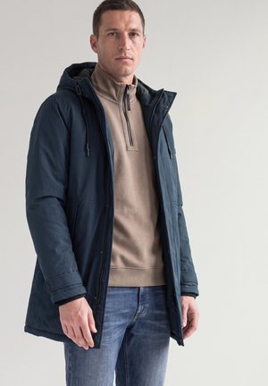 Homme portant une veste d'hiver à capuche bleu marine par-dessus un pull beige à fermeture éclair quart de zip et un jean bleu, debout devant un fond clair uni.