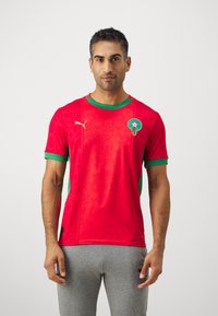 Maglietta a maniche corte rossa con motivo di stelle, dettagli verdi e colletto tondo. Include un logo sul lato sinistro del petto.