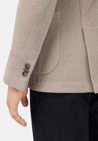 Beige stickad kavaj med strukturerad tyg, en ficka och manschetter med tre knappar, i kombination med mörka byxor. Fokusera på ärmdetaljen och passformen.