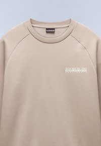 Suéter beige con cuello redondo y mangas raglán. Presenta una superficie texturizada y un logo impreso en blanco en el lado izquierdo del pecho. Tejido suave.