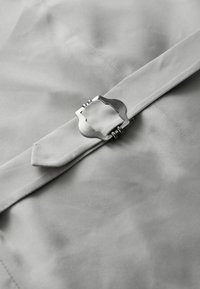 Pilkas audinys su lygiu tekstūru, su sidabriniu metalo sagtimi ir smailu diržo detaliu, pabrėžiančiu minimalizmą.