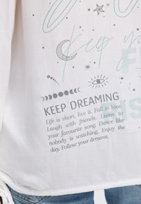 Weißer Stoff mit bedruckten Phrasen "KEEP DREAMING" und aufmunterndem Text, Mondphasen, Sterne, ein Auge und abstrakten türkisfarbenen Linienmustern.