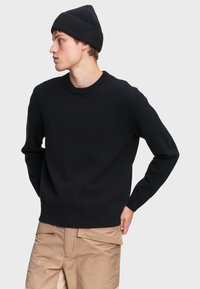 Čierny pletený sveter s okrúhlym výstrihom a dlhými rukávmi, doplnený zodpovedajúcou rebrovanou čiapkou, nosený s béžovými cargo nohavicami.