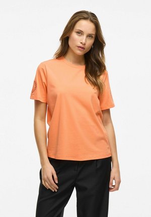 Camiseta de manga corta en algodón naranja brillante, con un cuello redondo y un diseño decorativo con recortes en la manga izquierda. Pantalones negros visibles.