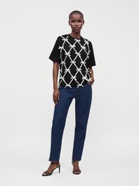 Femme portant un haut noir à manches courtes avec des détails de nœuds blancs, un jean bleu et des sandales à talons noires, debout devant un fond uni.