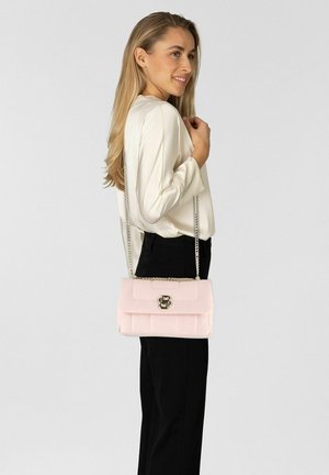 Frau trägt weiße Bluse und schwarze Hose, trägt eine hellrosa Umhängetasche mit goldener Kettenriemen und Logoverschluss.