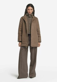 Brun knäppt jacka med hög krage, lager på lager över en ribbad polotröja. Matchad med bruna vida byxor, vilket skapar en monokromatisk look.