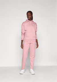 Roze hoodie en sweatpants set gemaakt van zachte stof, met een voorzak en logo, gecombineerd met witte sportschoenen.