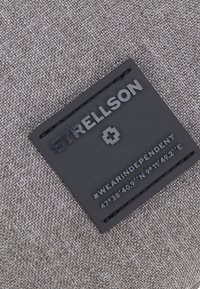 Černá obdélníková textilní etiketa s vyraženým textem "STRELLSON" a hashtagem "#WEARINDEPENDENT", s texturovaným povrchem, připevněná černým stehem.