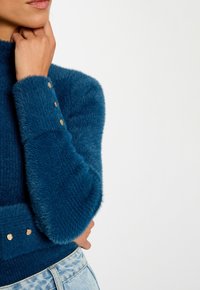 Magic sweater verde acqua con colletto alto e una texture a coste unica. Caratterizzato da bottoni dorati sui polsini e un dettaglio in vita aderente.
