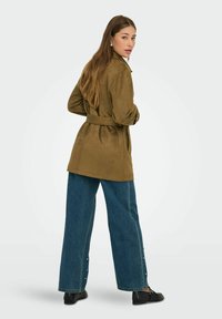 Olivgrön mockajacka med bälte och skräddarsydd passform, i kombination med vida blå jeans med sidoknappar och svarta skor.