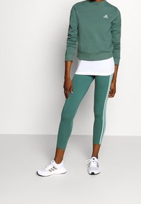 Sudadera verde de algodón con puños acanalados, combinada con leggings a juego que tienen franjas laterales blancas y zapatillas deportivas blancas.