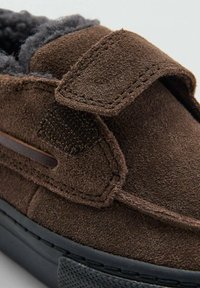 Zapato marrón de ante tipo slip-on con un forro gris peludo, correa ajustable de velcro y una suela de goma negra con acentos texturizados.