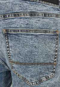 Un tissu de denim délavé clair présente une seule poche arrière, soulignée de surpiqûres orange et d'un léger détail textural à la surface.