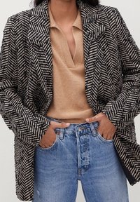 Marynarka w wzór houndstooth w czarno-białym kolorze, z wyciętym kołnierzem i teksturowanym materiałem. Połączona z beżową bluzką i jasnoniebieskimi dżinsami.