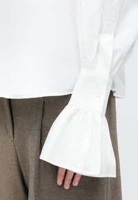 Camicia bianca in cotone con maniche ampie e plissettate, dotata di polsini con bottoni, abbinata a pantaloni marroni strutturati. Vista ravvicinata dei dettagli delle maniche e dei polsini.