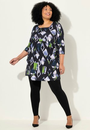 Une tunique noire avec un motif géométrique bleu et vert, présentant un décolleté carré et des manches ¾, associée à des leggings noirs et des talons.