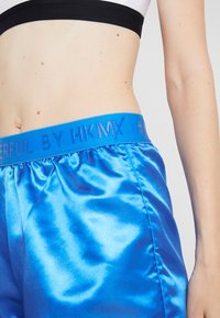 Satin blue shorts with an elastic waistband featuring "POWERFUL BY HKMX" text in a contrasting blue font. Smooth, shiny texture.