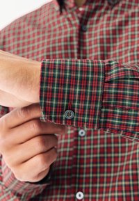 Camicia a quadri rossa e verde con polsini con bottoni, caratterizzata da un tessuto strutturato e cuciture visibili. Primo piano di una mano che sistema il polsino.