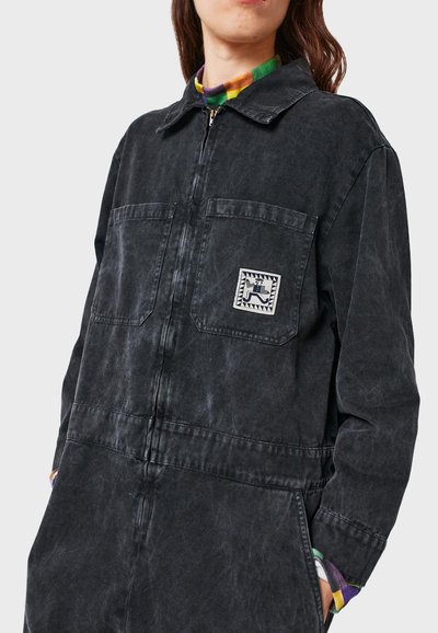 Schwarzer Denim-Overall mit einem Frontreißverschluss, zwei Brusttaschen und einem charakteristischen Logo-Patch. Verfügt über einen lockeren Schnitt und eine subtile, ausgebleichte Textur.