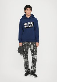 Felpa blu navy con logo "VERSACE COUTURE", abbinata a pantaloni con fantasia bianca e nera, e un cappellino grigio in mano. Sneakers nere.