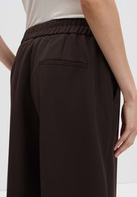 Personne portant un pantalon élastique marron foncé avec une poche passepoilée au dos, et un haut blanc, se tenant contre un fond uni.