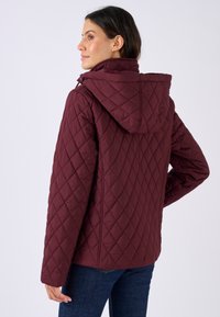 Giacca trapuntata color bordeaux con cappuccio e tasche laterali. Presenta una superficie testurizzata e una vestibilità comoda, abbinata a jeans blu.