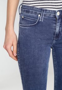 Närbild av en person som bär blå denimjeans och en randig skjorta, hand avslappnad vid sidan.
