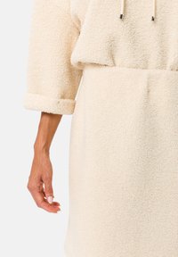 Abito in morbido tessuto di colore crema con una vestibilità rilassata, cappuccio oversize e maniche arrotolate. Superficie testurizzata con fibre visibili.