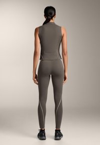 Vrouw die met haar rug naar de camera staat, gekleed in een mouwloos nauwsluitend topje en bijpassende leggings met reflecterende zijstrepen, zwarte sneakers, neutrale achtergrond.