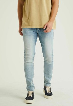 Mężczyzna w jasnoniebieskich jeansach typu slim-fit, beżowej koszulce i czarnych trampkach z białymi podeszwami, stojący na jasnym tle.