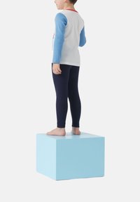 Bambino che indossa una maglietta a maniche lunghe con maniche blu e dettagli rossi, abbinata a leggings blu navy, in piedi su un cubo azzurro chiaro.