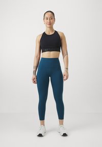 Top sportivo nero con cinturino "BEAT BETTER", leggings teal a vita alta, texture liscia, design aderente e scarpe da ginnastica bianche.