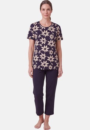 Vrouw staat blootsvoets en draagt een marineblauw gebloemd shirt met korte mouwen en marineblauwe cropped broek tegen een effen achtergrond.