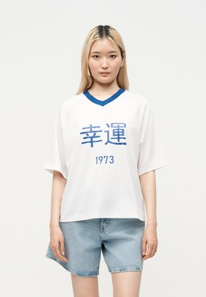 Jonge vrouw met recht blond haar die een wit V-hals shirt draagt met blauwe rand en blauwe tekens en spijkershorts, staand tegen een effen achtergrond.