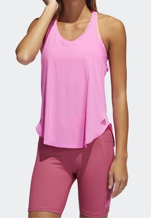 Femme portant un débardeur rose clair et un short de sport ajusté rose foncé, debout avec une main près du visage et l'autre détendue.