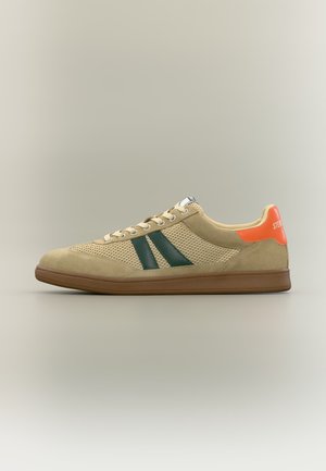 Nizka bež športna copatka z mrežastimi stranicami, temno zelenimi črtami na strani, rjavim podplatom in oranžnim jezičkom na peti z belim napisom.