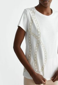 T-shirt bianco in cotone a maniche corte con un motivo a catena dorata creato con decorazioni lungo un lato.