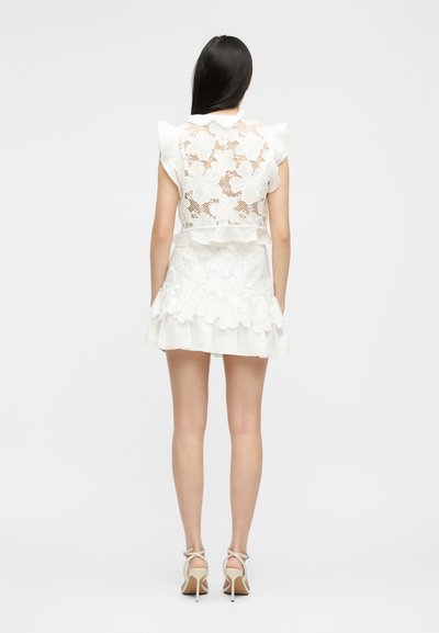 Γυναίκα με μακριά σκουρόχρωμα μαλλιά φοράει λευκό τοπ από δαντέλα με λουλούδια και ταιριαστή φούστα με βολάν, συνδυασμένα με λευκές ψηλοτάκουνες σανδάλια, γυρισμένη προς τα πίσω.