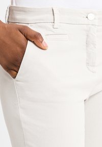 Hellgraue Baumwollhose mit glatter Textur, ausgestattet mit Fronttaschen und einem einfachen Knopfverschluss. Eine Tasche hat eine gestickte Akzentuierung.