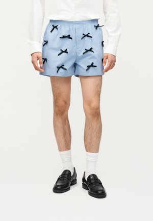 Άνδρας που φοράει ανοιχτό μπλε σορτς με πολλούς μικρούς μαύρους φιόγκους, λευκές κάλτσες, μαύρα λουστρίν παπούτσια και λευκό πουκάμισο μακρυμάνικο, στέκεται με τα χέρια χαλαρά.